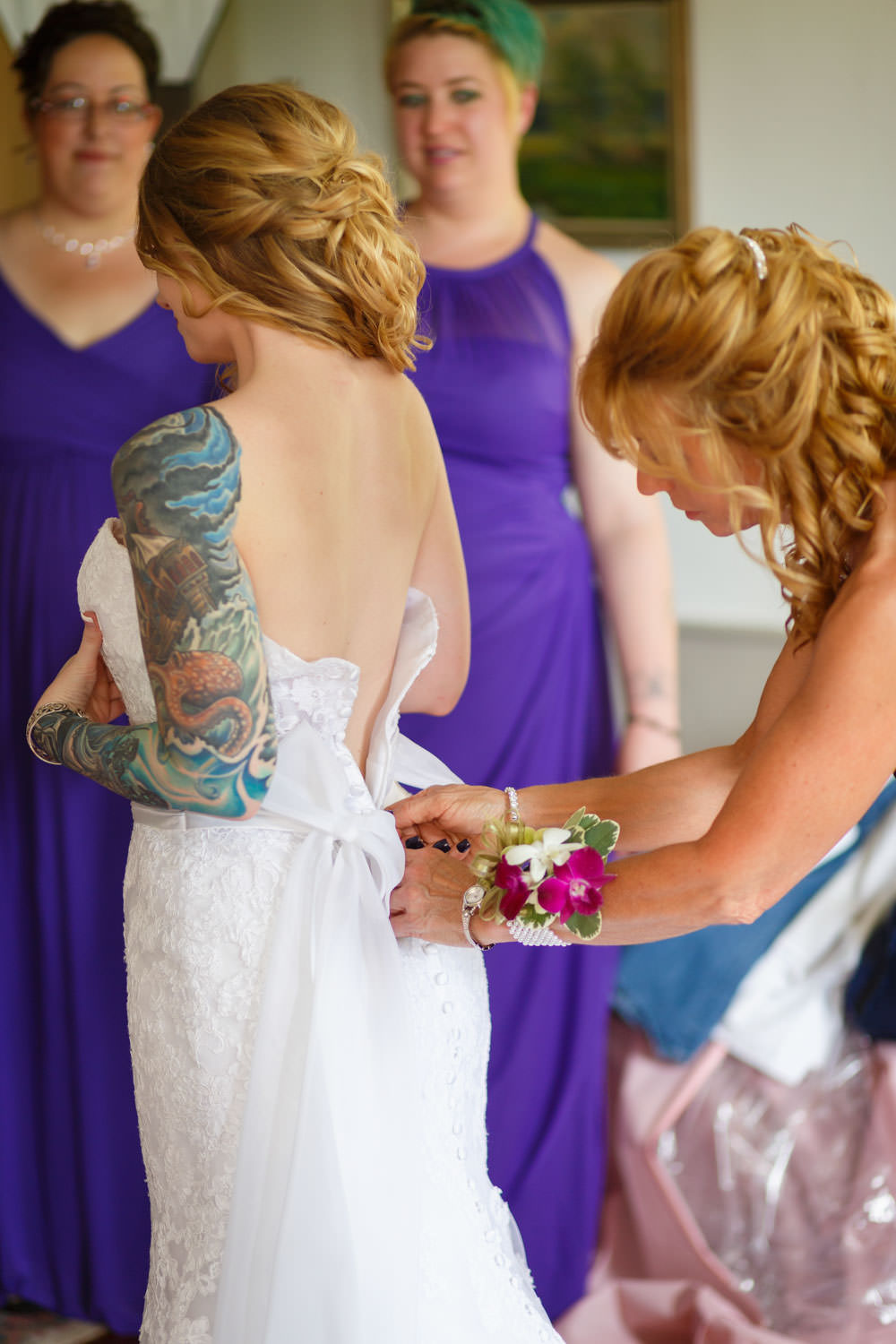 The brides mother zipping up her wedding dress. Purple bridesmaids dresses in the background in the bridal suite. The bride has intricate tattoo on her left arm.