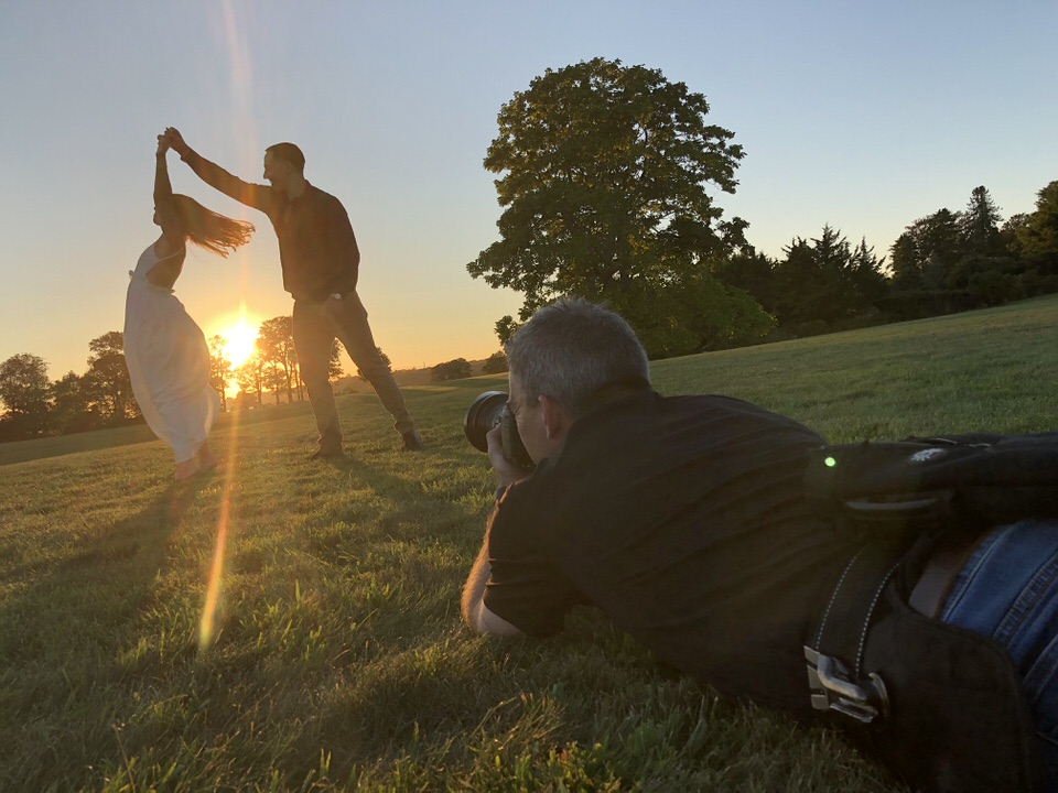 Photographer laying on the ground taking sunset photo of couple while guy twirls girl