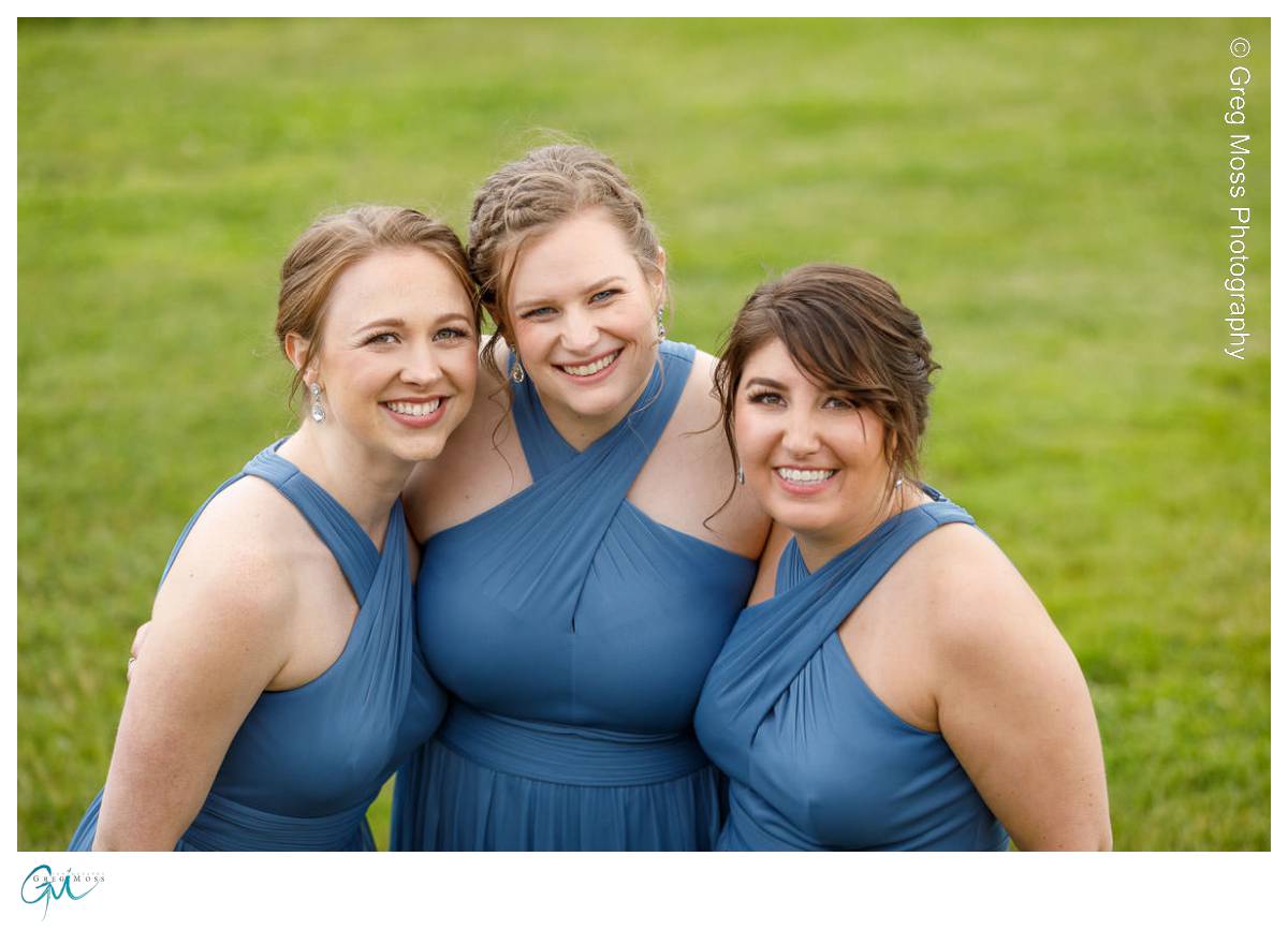 Red Barn Wedding0543 Three women in matching blue dresses smiling together at an outdoor wedding, with a red barn and soft-focus green grass background.