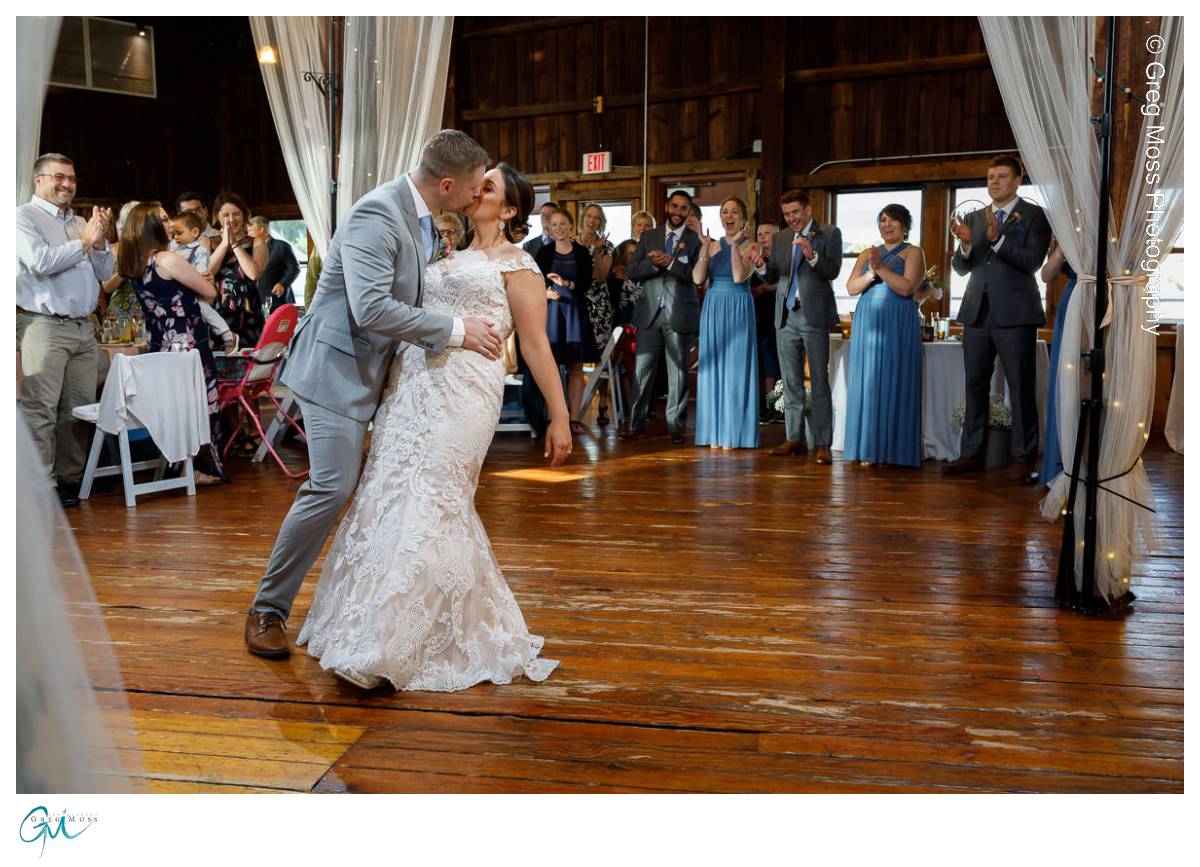 Red Barn Wedding0544 A newlywed couple sharing a kiss during their first dance at a red barn wedding venue, surrounded by applauding guests.
