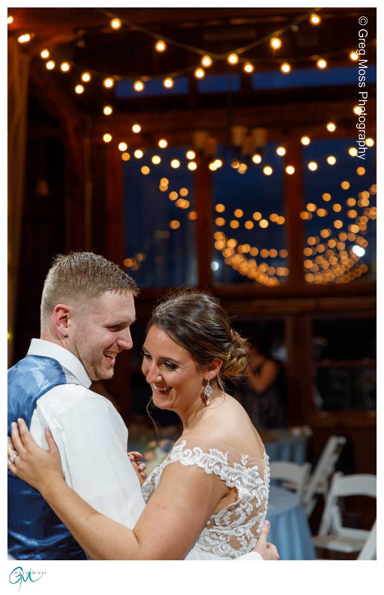 Red Barn Wedding0545 A bride and groom share a close dance under string lights in a rustic red barn, smiling warmly at each other.