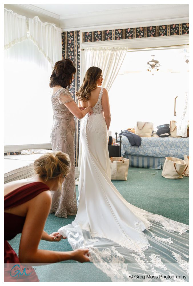 Maid of honor fluffing dress while mother of the bride buttons the brides dress