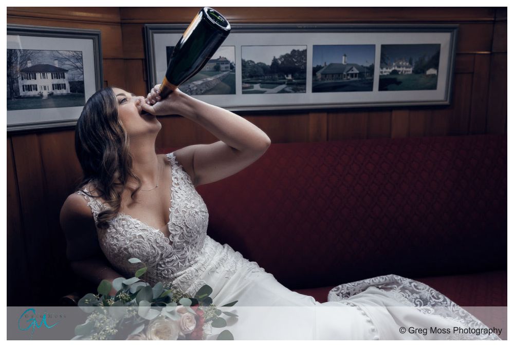 Bride laying on red couch with bridal bouquet while jugging bottle of champagne