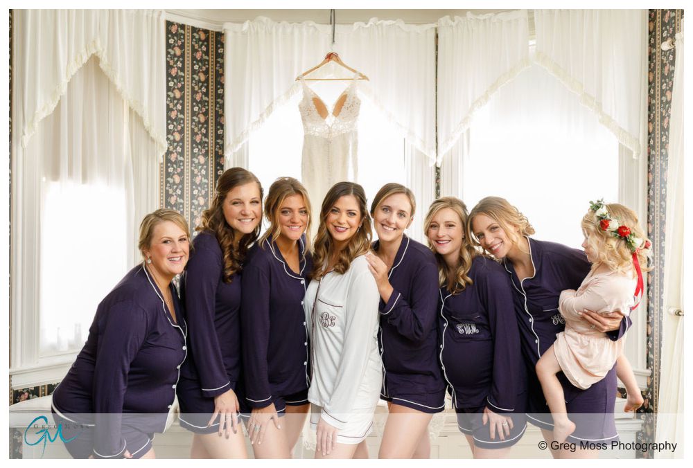 Bride in white robe and bridesmaids in blue pajamas with the brides dressing hanging in the background