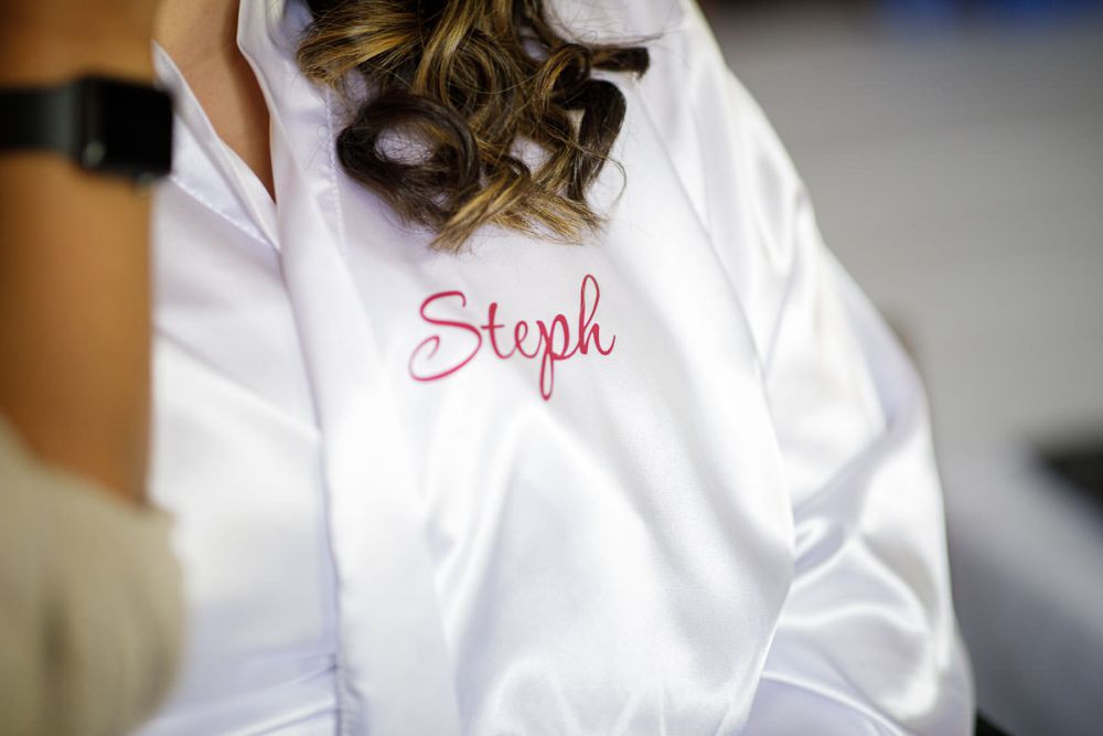 Wrights Mill Farm Wedding-2 Close-up of a white robe with the name "Steph" embroidered in pink, worn by a woman with curly hair at a Wrights Mill Farm Wedding.