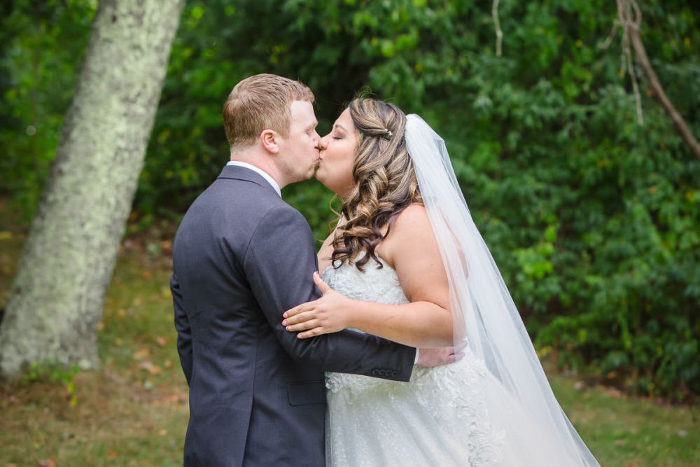Wrights Mill Farm Wedding-8 A bride and groom kissing outdoors at Wrights Mill Farm, surrounded by green foliage. The bride is wearing a white dress and veil, and the groom is in a dark suit.