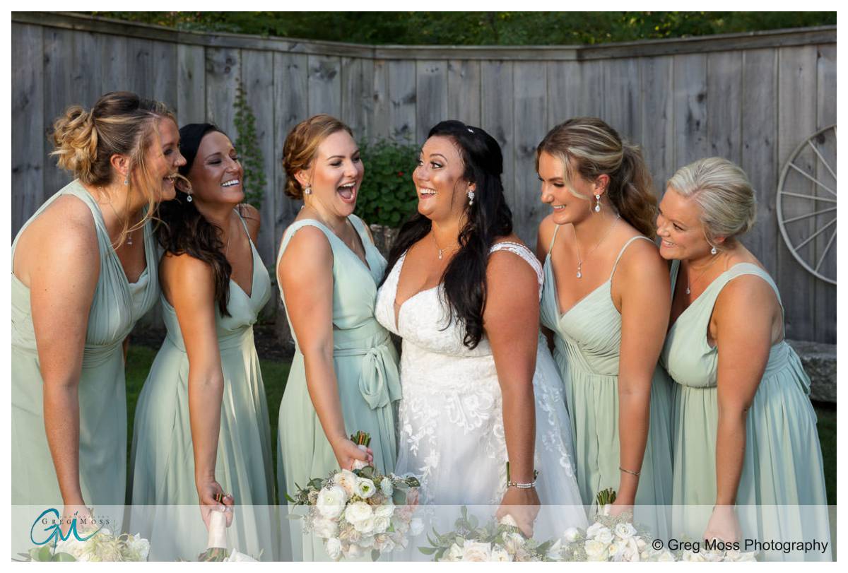 Blissful Meadows G. C. Wedding011 A bride laughing joyfully surrounded by five bridesmaids in matching light green dresses at Blissful Meadows Wedding, with wooden fence and lush greenery.