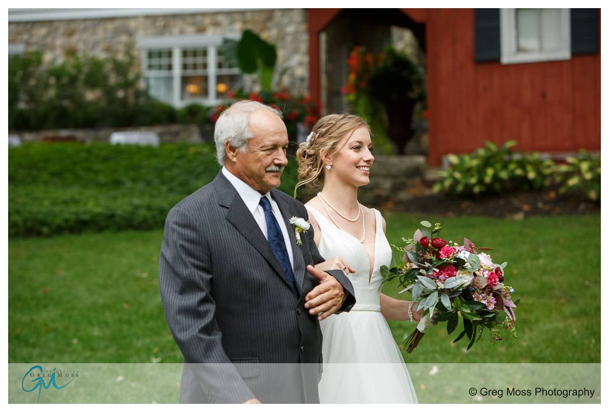 Publick house vov renewal 013 A bride in a white dress, holding a bouquet, walks arm-in-arm with an older man wearing a suit, in front of the Publick House.