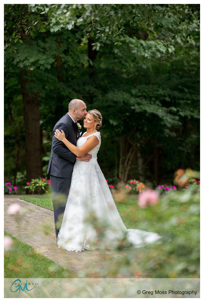 Log Cabin Holyoke Weddings-12 A bride and groom embrace and laugh together amidst lush greenery and vibrant flowers at their Holyoke log cabin wedding.