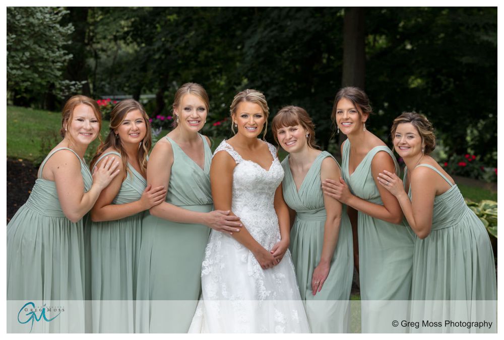 Log Cabin Holyoke Weddings-13 A bride in a white dress surrounded by six bridesmaids in matching light green dresses, posing outdoors near a log cabin in Holyoke.