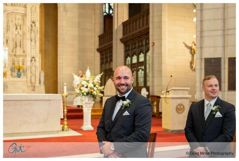 Log Cabin Holyoke Weddings-15 Two men in suits, one standing and smiling at the camera, the other seated and looking ahead, inside a log cabin with floral decorations.