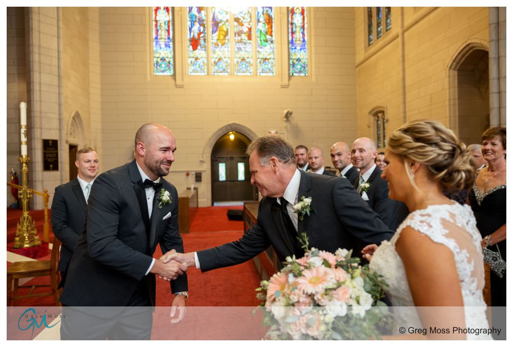 Log Cabin Holyoke Weddings-16 A bride and groom smiling at each other as the groom shakes hands with an older man at the altar of a church, with guests looking on during their Holyoke wedding.