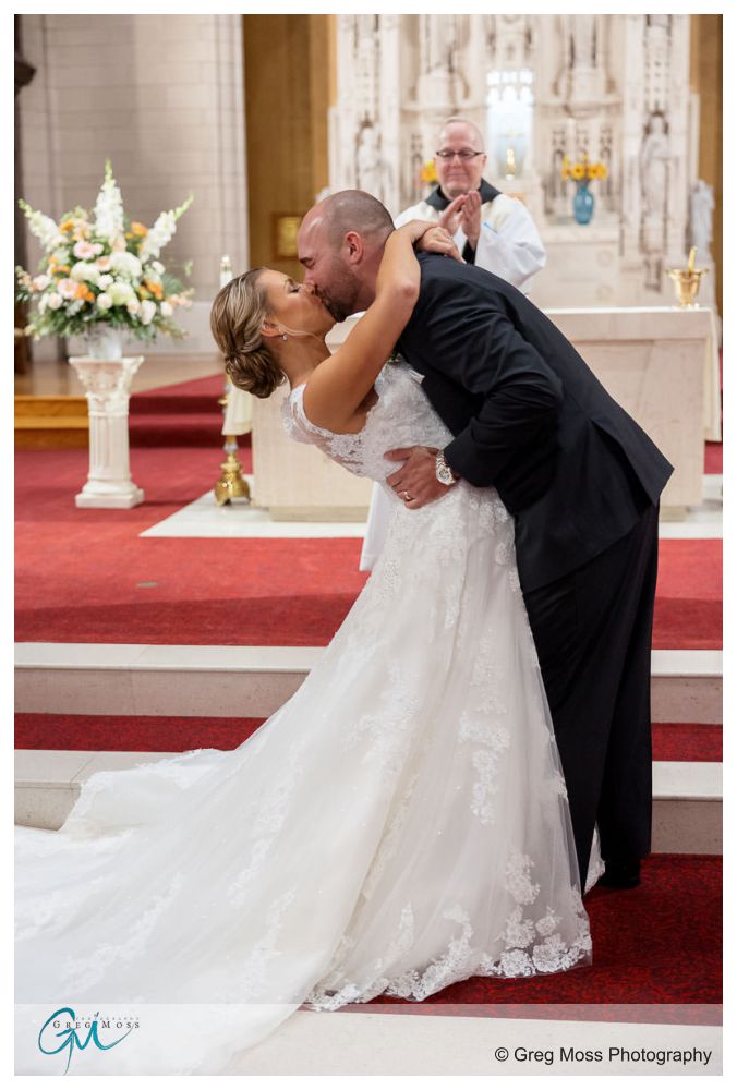 Log Cabin Holyoke Weddings-17 Bride and groom kissing passionately at the altar, with a priest in the background, in a log cabin decorated with flowers.