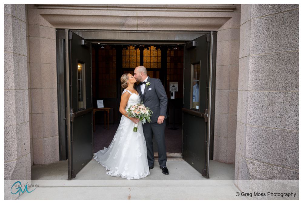 Log Cabin Holyoke Weddings-18 A bride and groom kiss in a doorway, framed by stone walls, with the groom holding a bouquet during their Holyoke wedding.