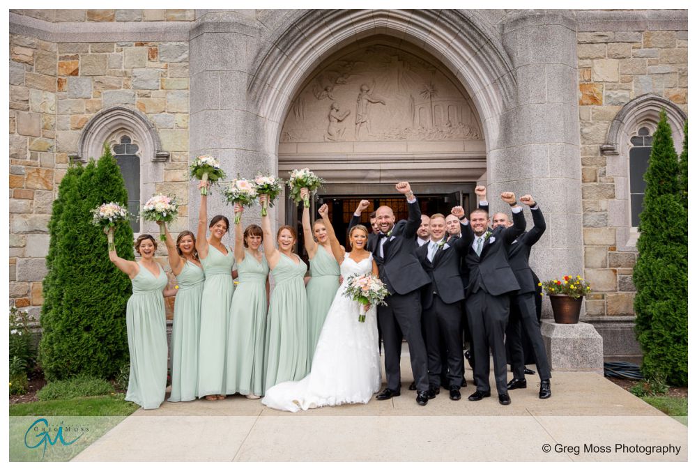 Log Cabin Holyoke Weddings-19 A joyful wedding group posing outside a log cabin, with bridesmaids in pale green dresses and groomsmen in black tuxedos, all cheering.