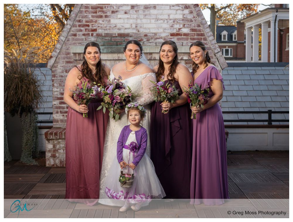 Inn on Boltwood Fall wedding-10 Four women and a young girl in coordinated purple dresses holding bouquets pose outdoors at Inn on Boltwood, with a brick backdrop ideal for a fall wedding.