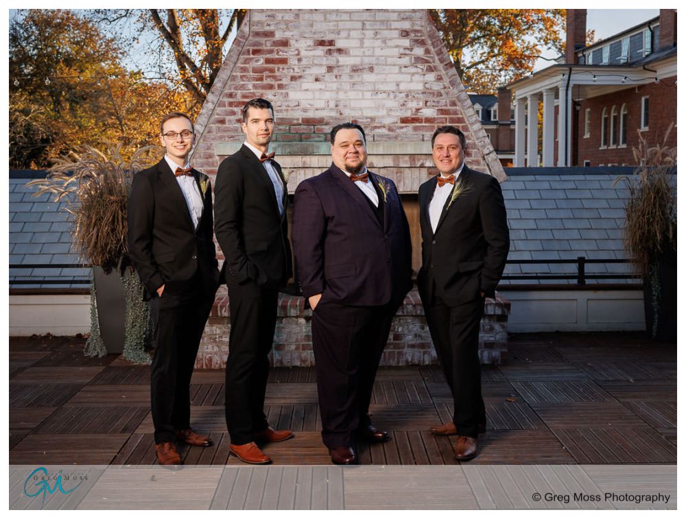 Inn on Boltwood Fall wedding-11 Four men in tuxedos standing confidently on a rooftop deck at an Inn on Boltwood fall wedding, with a brick structure and trees in the background.
