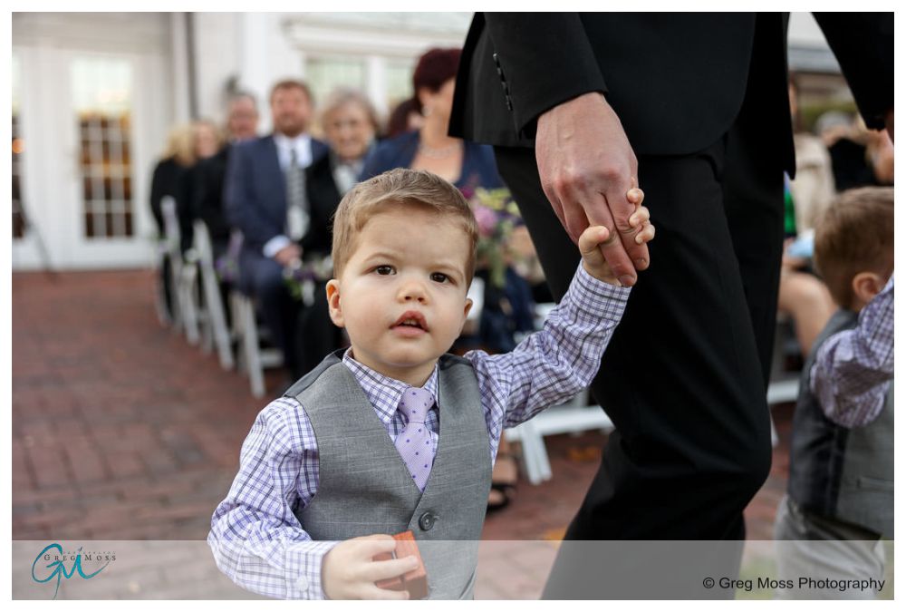 Inn on Boltwood Fall wedding-12 A young boy in a gray vest and purple tie looks up while holding an adult's hand at an Inn on Boltwood fall wedding, with guests in the background.
