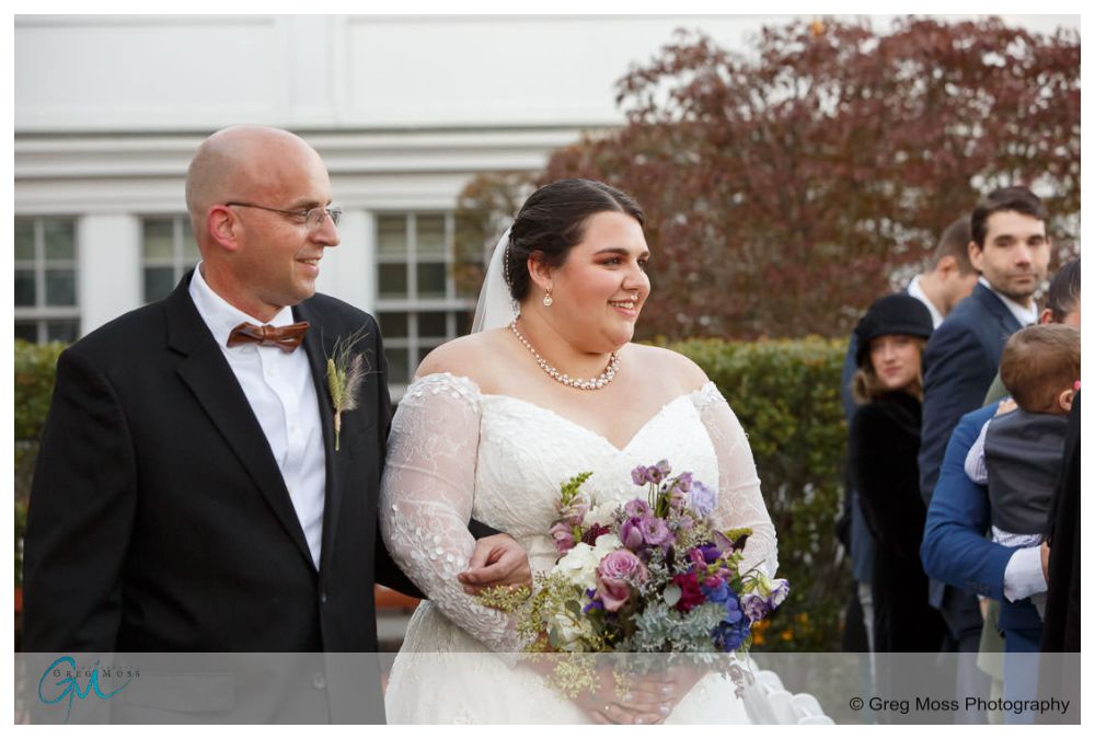Inn on Boltwood Fall wedding-13 A bride accompanied by her father walks joyfully at an outdoor fall wedding at the Inn on Boltwood, with guests in the background.