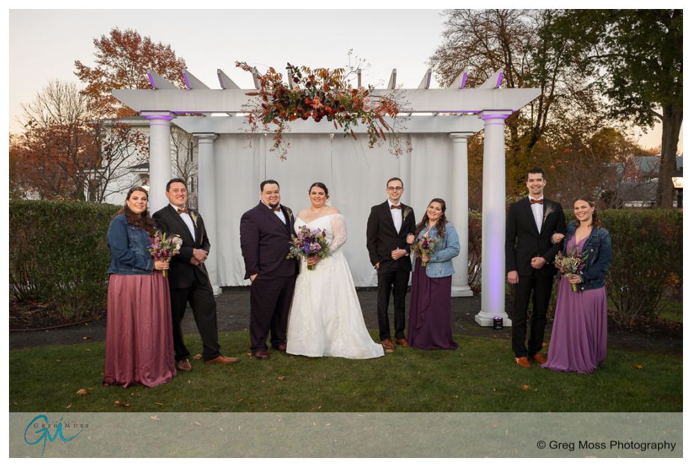 Inn on Boltwood Fall wedding-16 A wedding party posing in front of a white altar adorned with flowers at the Inn on Boltwood during a fall sunset.