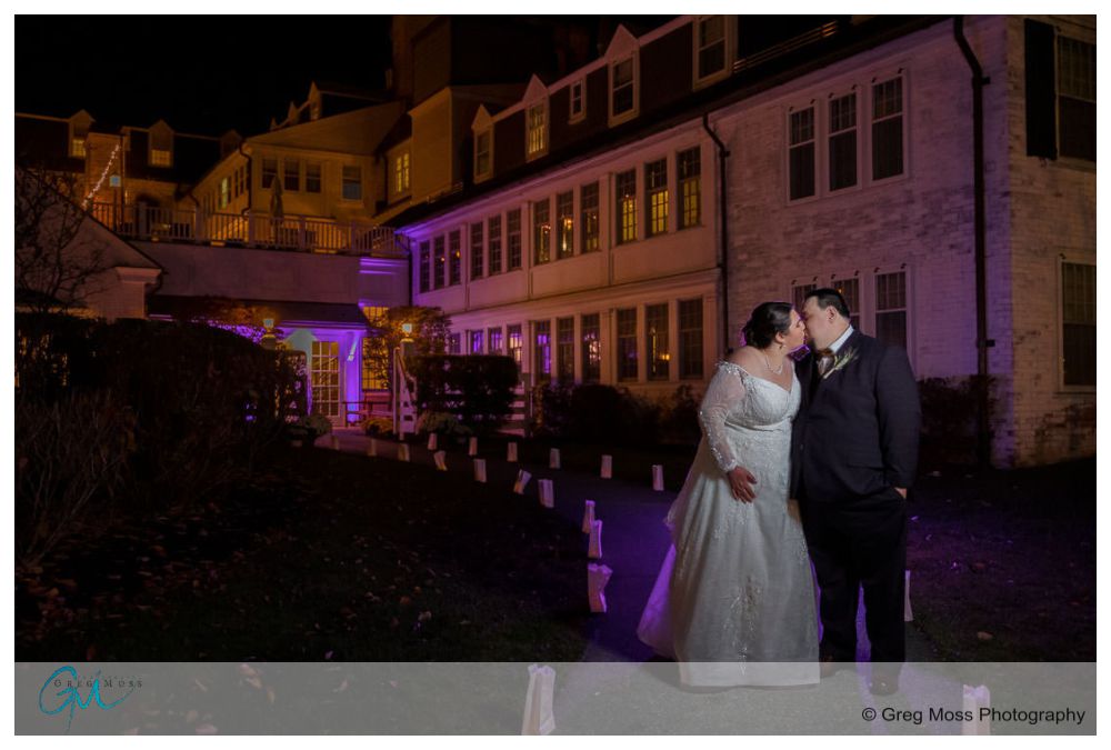 Inn on Boltwood Fall wedding-20 A couple in wedding attire kiss at night in front of the brightly lit, multi-story Inn on Boltwood with lawn and path luminaries.