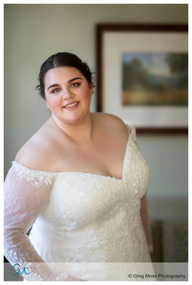 Inn on Boltwood Fall wedding-4 A bride in an off-the-shoulder lace wedding dress smiles gently, standing in a room with a landscape painting in the background at the Inn on Boltwood fall wedding.