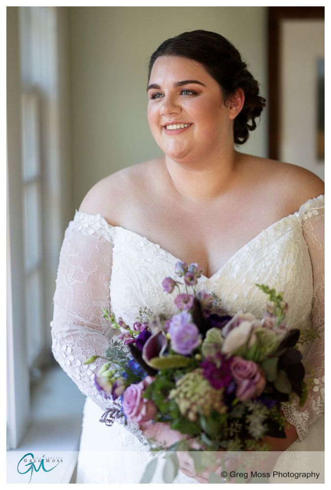 Inn on Boltwood Fall wedding-6 A bride smiling in a lace wedding dress, holding a bouquet of purple and green flowers, standing by a window at the Inn on Boltwood during a fall wedding.