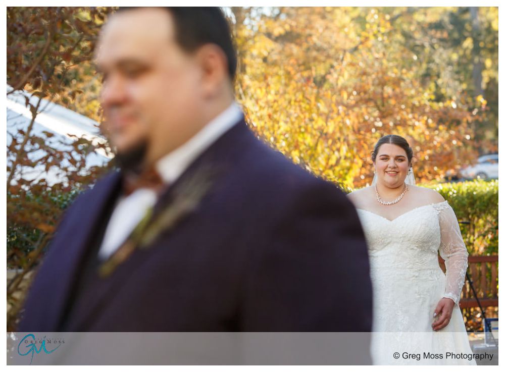 Inn on Boltwood Fall wedding-8 A groom in focus in the foreground with a smiling bride approaching from behind at an Inn on Boltwood fall wedding in a sunlit outdoor setting.
