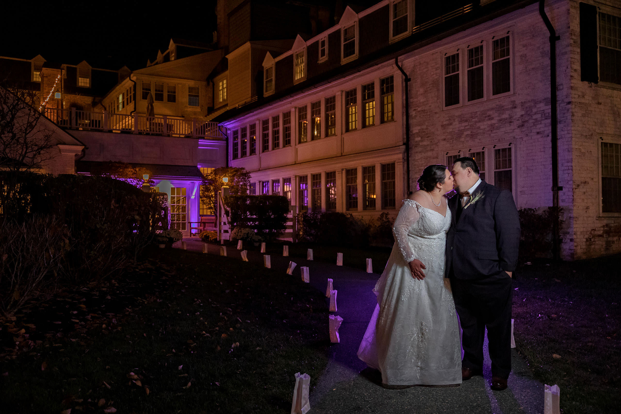 A couple in formal attire sharing a kiss at night outside a lit-up building with purple lighting during their fall wedding at the Inn on Boltwood.