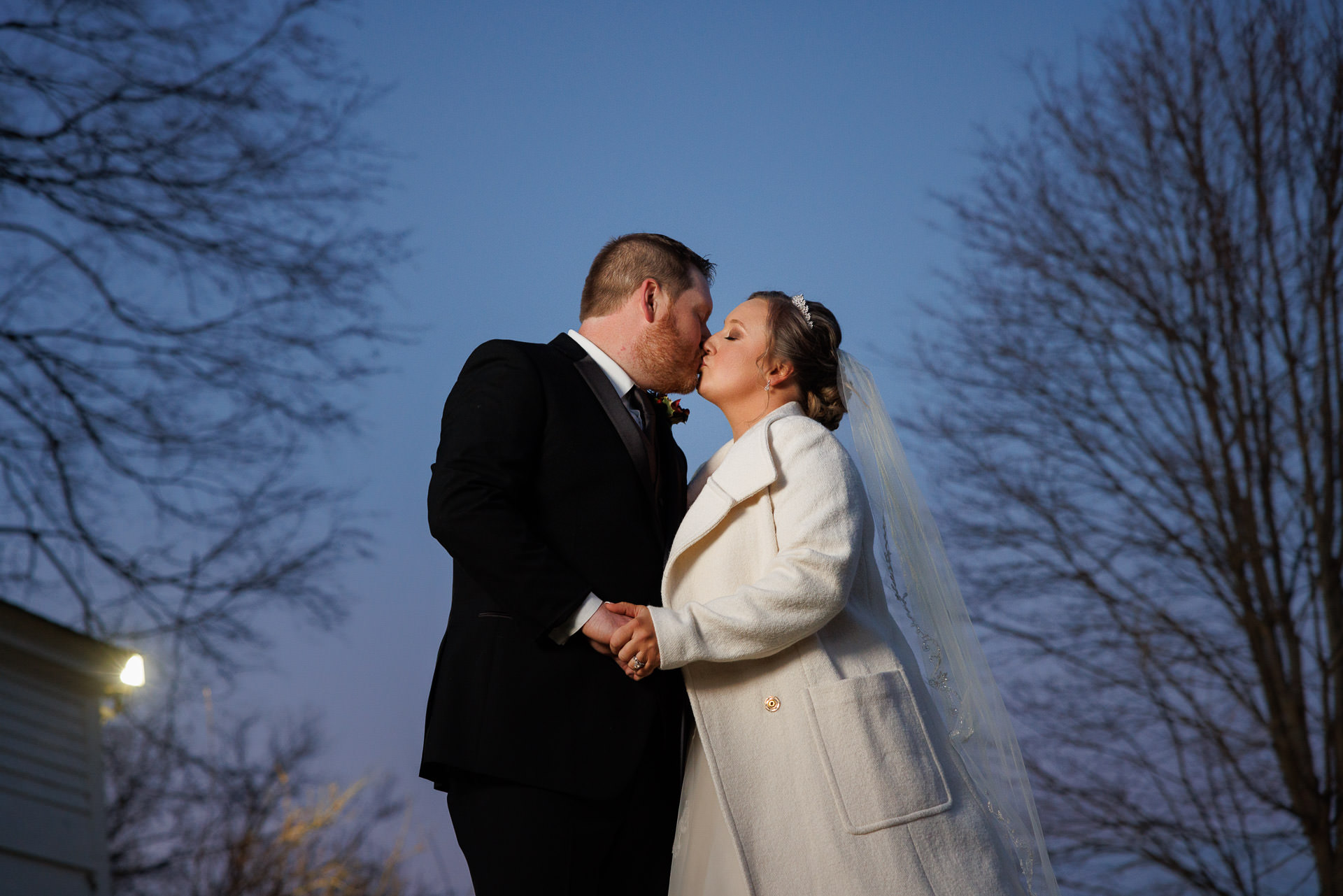 A bride in a white coat and veil and a groom in a black suit kissing outdoors at dusk under a clear sky at their wedding at the Salem Cross Inn.