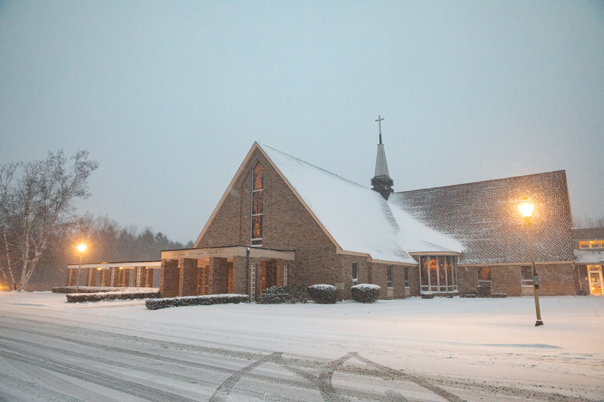 A snow-covered church with lit windows during a heavy snowfall at twilight in Holyoke, MA, with tire tracks on the driveway.