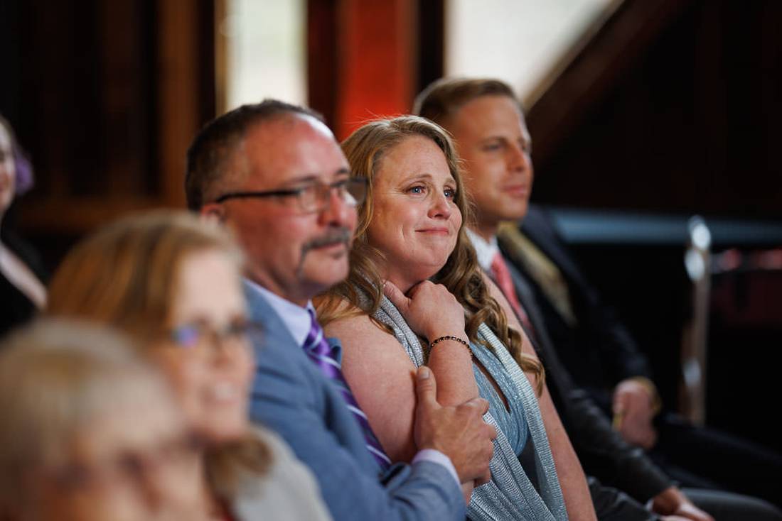 Red Barn Wedding Photography-12 Audience members seated in a row at a red barn wedding, focused intently on the event, with a woman in a blue dress looking emotional.