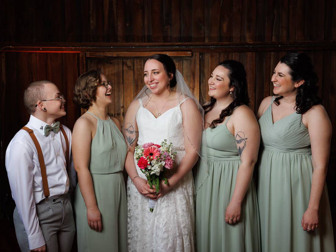 Red Barn Wedding Photography-16 A bride smiling in a white dress, holding a bouquet, stands with four attendants in matching sage dresses and tattoos inside a rustic red barn venue.