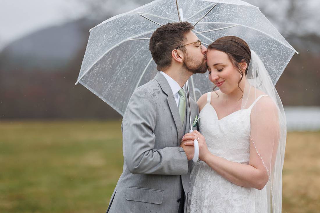 Red Barn Wedding Photography-18 A newlywed couple embracing under a transparent umbrella, the groom kissing the bride's cheek, with a misty landscape and a red barn in the background.