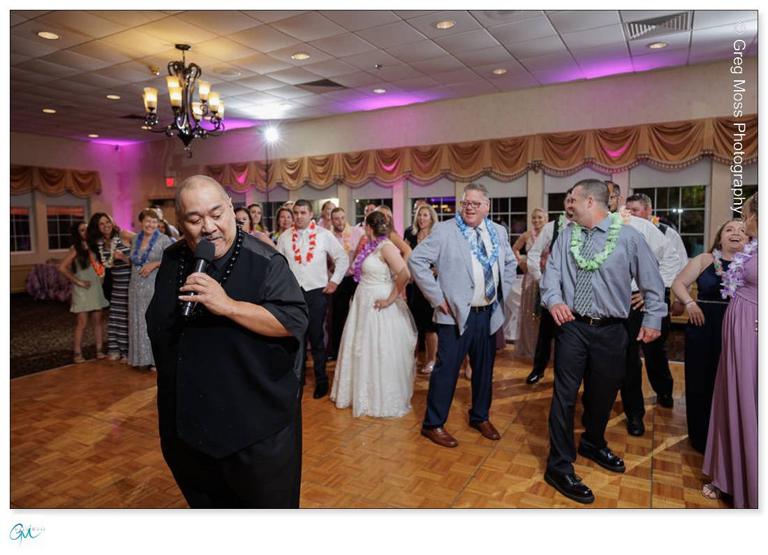 Holyoke Wedding Photography-20 A man sings into a microphone at a festive wedding event in Holyoke while a crowd in formal wear watches, some with leis around their necks, in a warmly lit banquet hall.