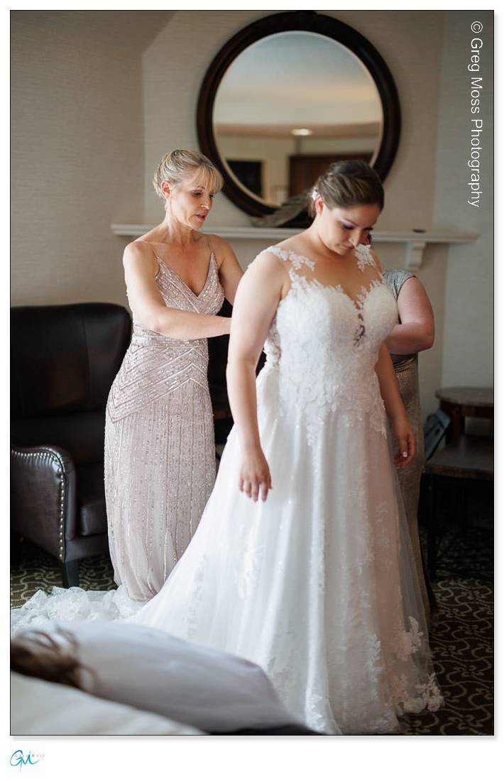 Holyoke Wedding Photography-3 A woman in a silver dress assists another woman in a white bridal gown, adjusting the gown's fit in a well-lit room during a Holyoke wedding photography session.