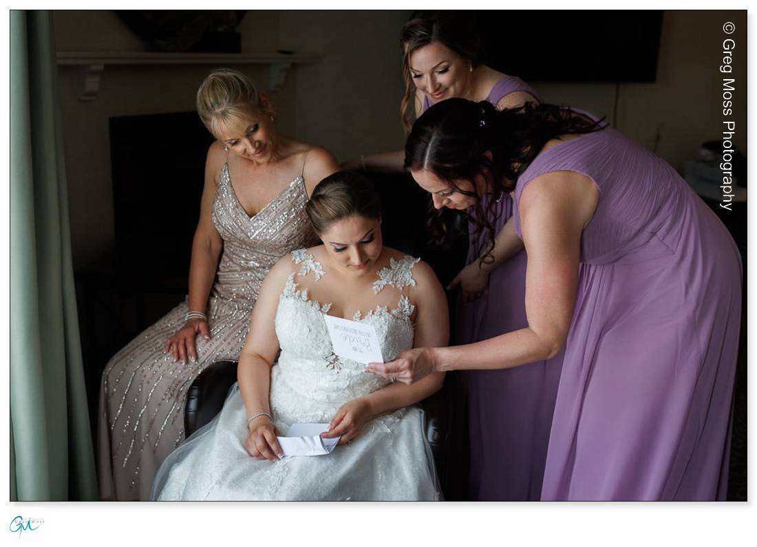 Holyoke Wedding Photography-5 A bride in a white gown reads a letter while surrounded by three women in elegant dresses, expressing emotional support at a Holyoke Wedding Photography session.