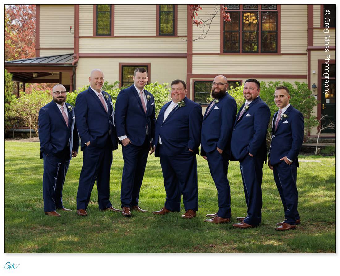 Highfield Hall Wedding-10 Group of seven men in matching navy blue suits and floral ties, posing outdoors in front of a two-story house at a Highfield Hall Wedding.