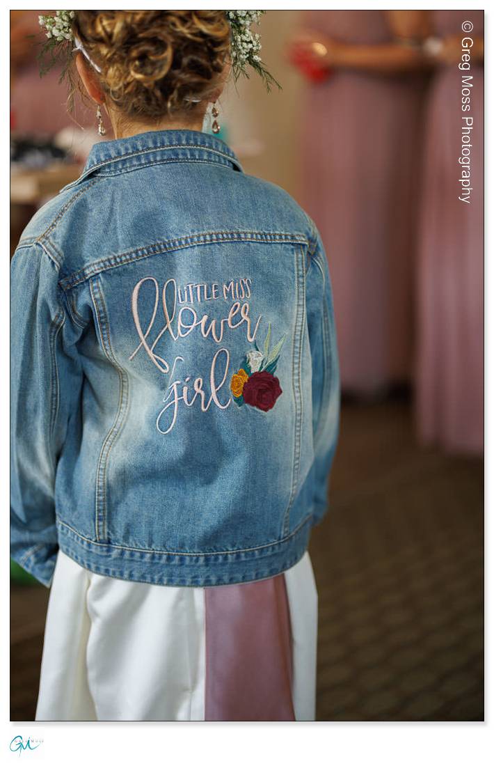 Highfield Hall Wedding-2 Young girl in a white dress wearing a denim jacket with "Highfield Hall flower girl" embroidered on the back, viewed from behind.