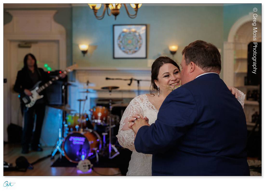Highfield Hall Wedding-25 A bride and groom share a dance, smiling at each other in a decorated room at Highfield Hall Wedding, with a live band performing in the background.