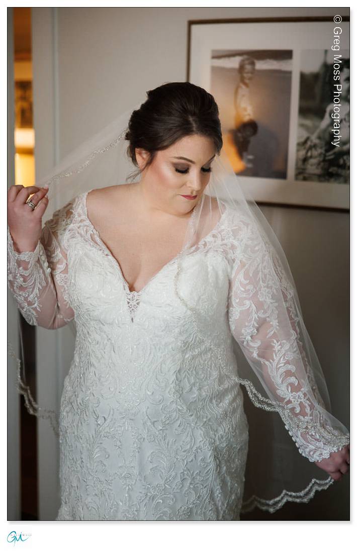 Highfield Hall Wedding-4 A bride in a lace wedding gown adjusts her veil indoors at Highfield Hall, softly lit, with a framed photo in the background.