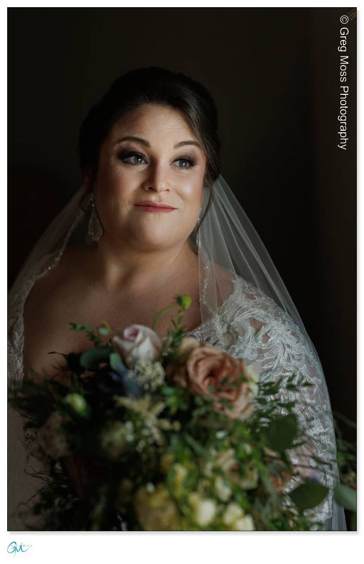 Highfield Hall Wedding-5 A bride in a lace dress and veil holds a bouquet at a Highfield Hall wedding, looking contemplatively to the side in soft lighting.