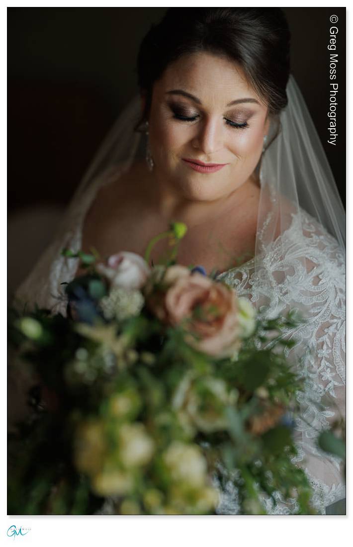 Highfield Hall Wedding-6 A bride in a lace gown and veil at a Highfield Hall wedding gazes downward at her bouquet, featuring roses in soft, pastel colors.