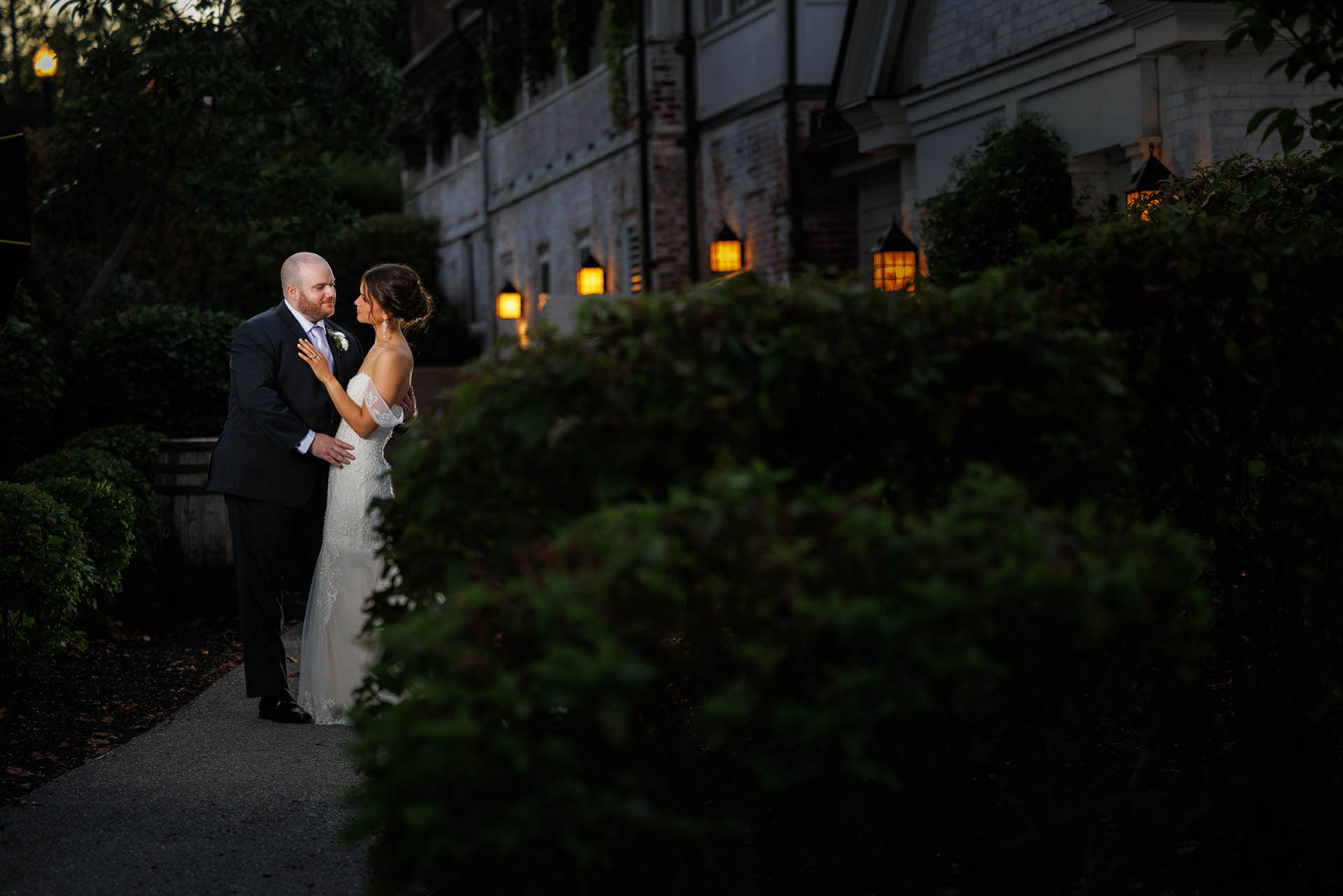 A couple in wedding attire embracing on a path near the Inn on Boltwood with warm lighting at dusk.