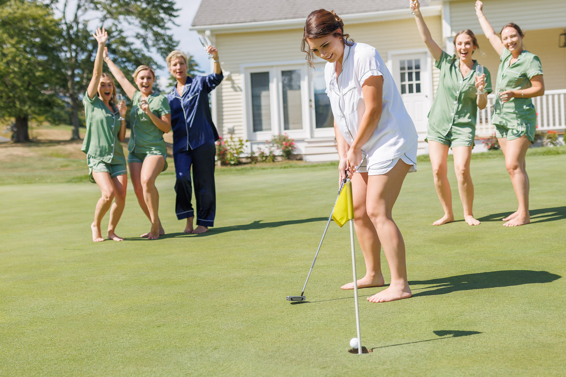 A woman in a white outfit putts a golf ball on a green with four cheering women in matching green outfits at The Ranch Wedding Photos event.