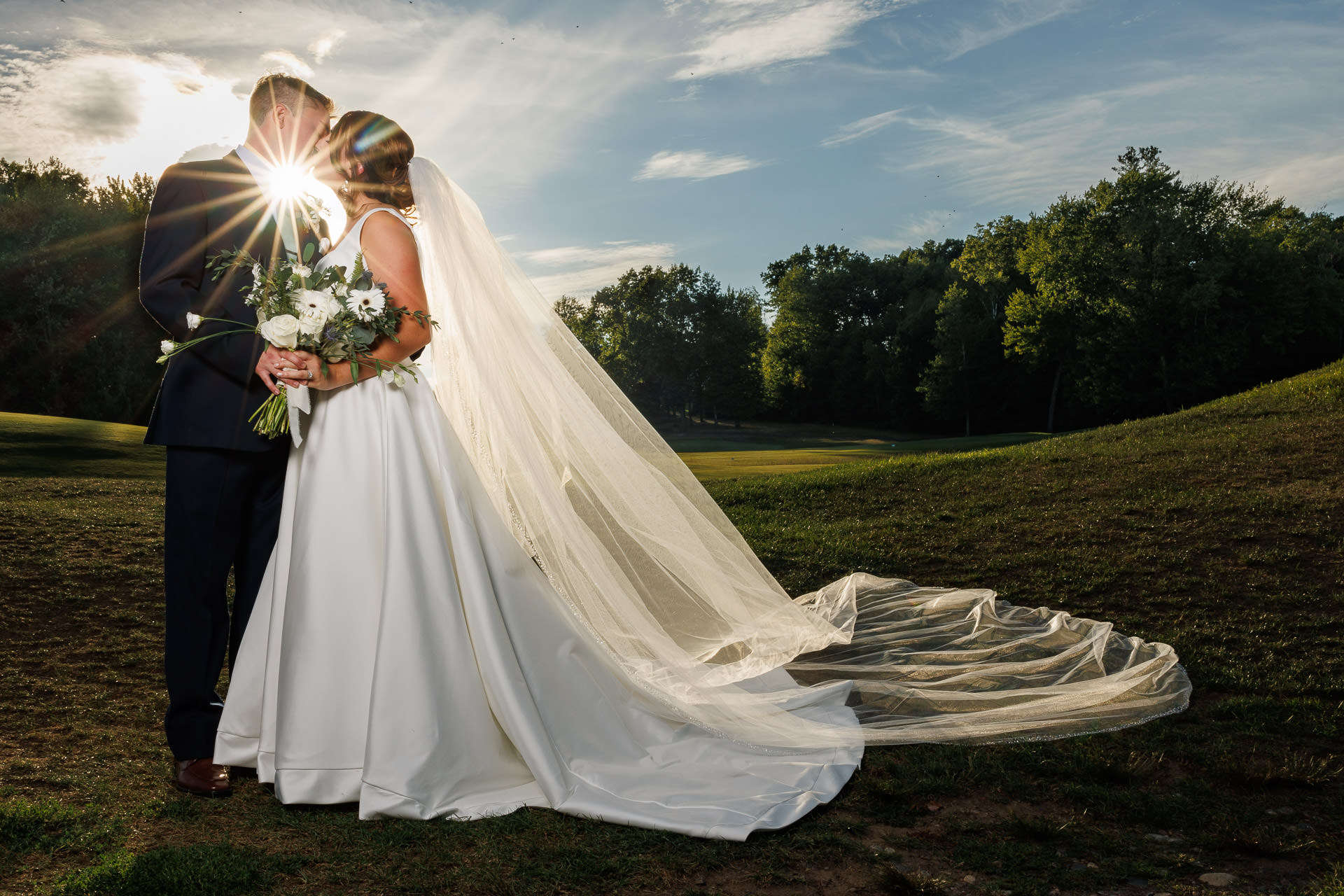 A bride and groom embracing outdoors at the ranch, with the sun shining through their kiss, highlighting the bride's flowing veil and the groom's dark suit.
