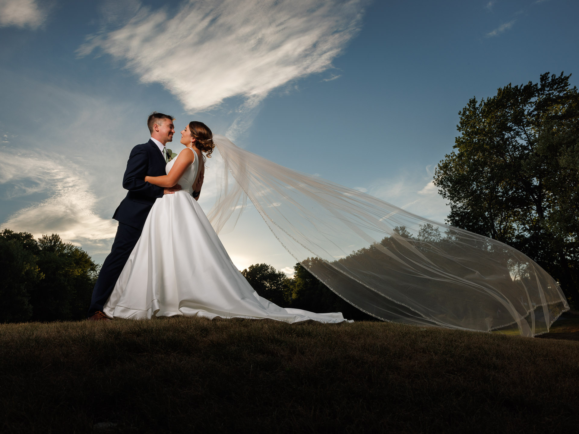 A bride and groom stand on a grassy field at The Ranch, the bride's long veil blowing in the wind against a dramatic sky at sunset.