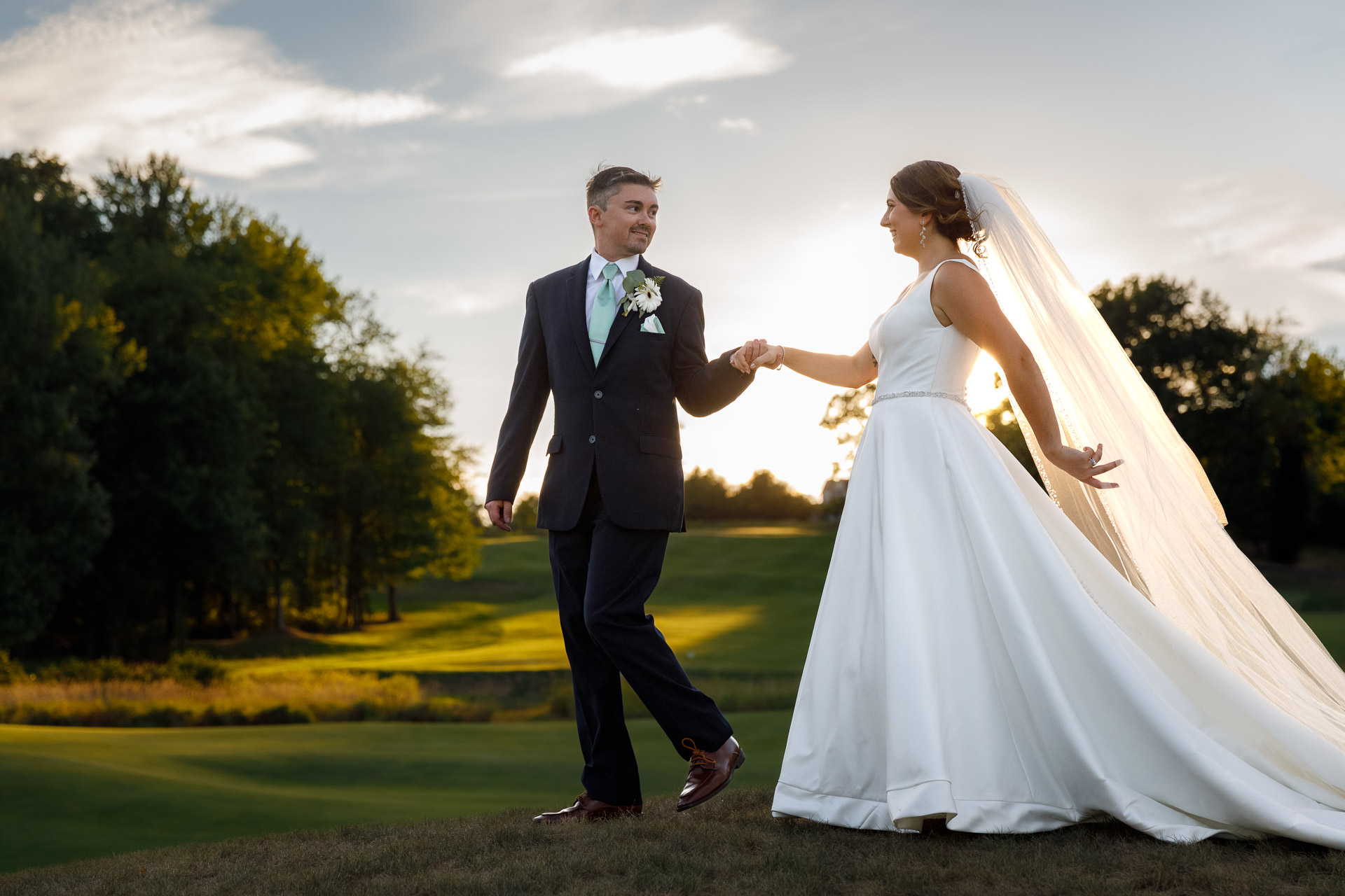 A bride and groom holding hands and walking across a grassy field at sunset at The Ranch, with trees and a clear sky in the background.