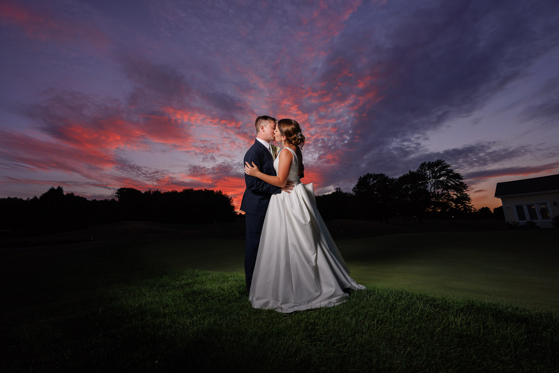 A bride and groom embracing and kissing on a golf course at sunset, with dramatic pink and blue clouds in the sky above during their Ranch wedding photoshoot.