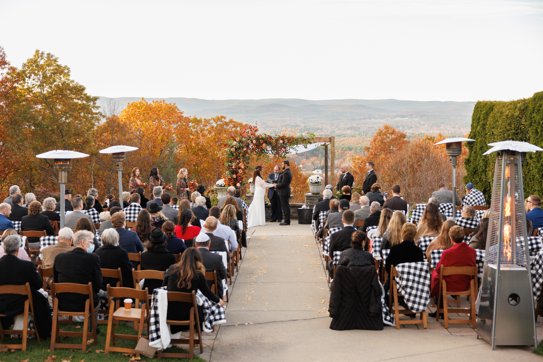 488 Lauren and William-1 An outdoor wedding ceremony with guests seated facing a couple at the altar, set against a backdrop of autumn hills, perfectly planned for exquisite wedding photography.
